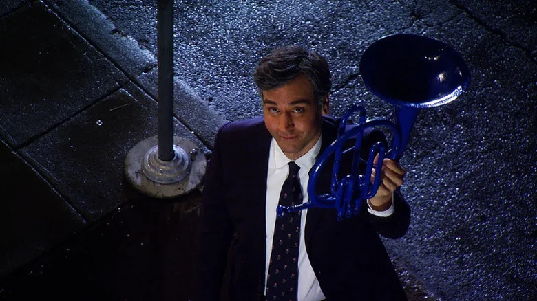 An older Ted Mosby (Josh Radnor) holds up a blue French horn while looking up from a sidewalk on How I Met Your Mother